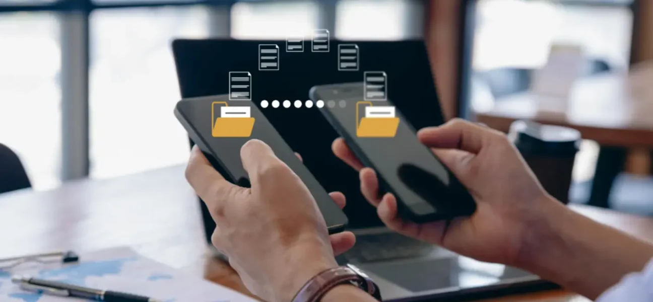 Oseba drži dva pametna telefona in prenaša podatke med njima. Na zaslonih sta prikazani ikoni map s puščicami, ki simbolizirajo prenos datotek. V ozadju je prenosni računalnik in kavarniško okolje