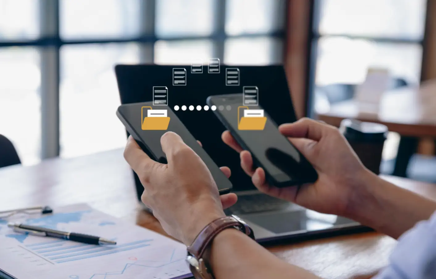 Oseba drži dva pametna telefona in prenaša podatke med njima. Na zaslonih sta prikazani ikoni map s puščicami, ki simbolizirajo prenos datotek. V ozadju je prenosni računalnik in kavarniško okolje