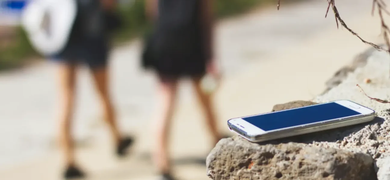 Pozabljen pametni telefon na kamnu ob poti, v ozadju se oddaljujeta dve osebi s nahrbtniki