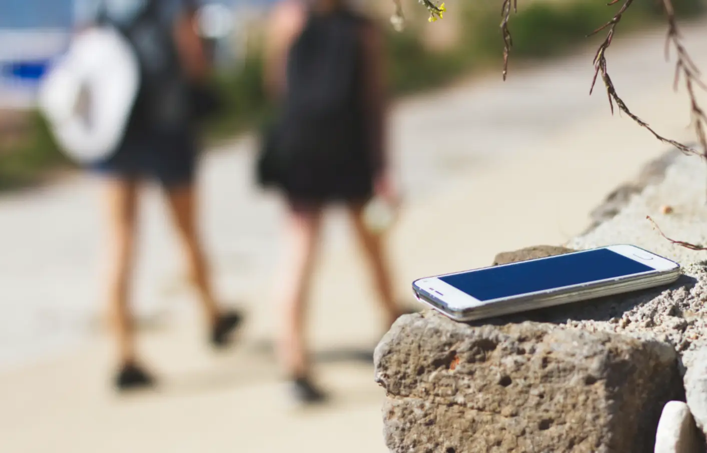 Pozabljen pametni telefon na kamnu ob poti, v ozadju se oddaljujeta dve osebi s nahrbtniki
