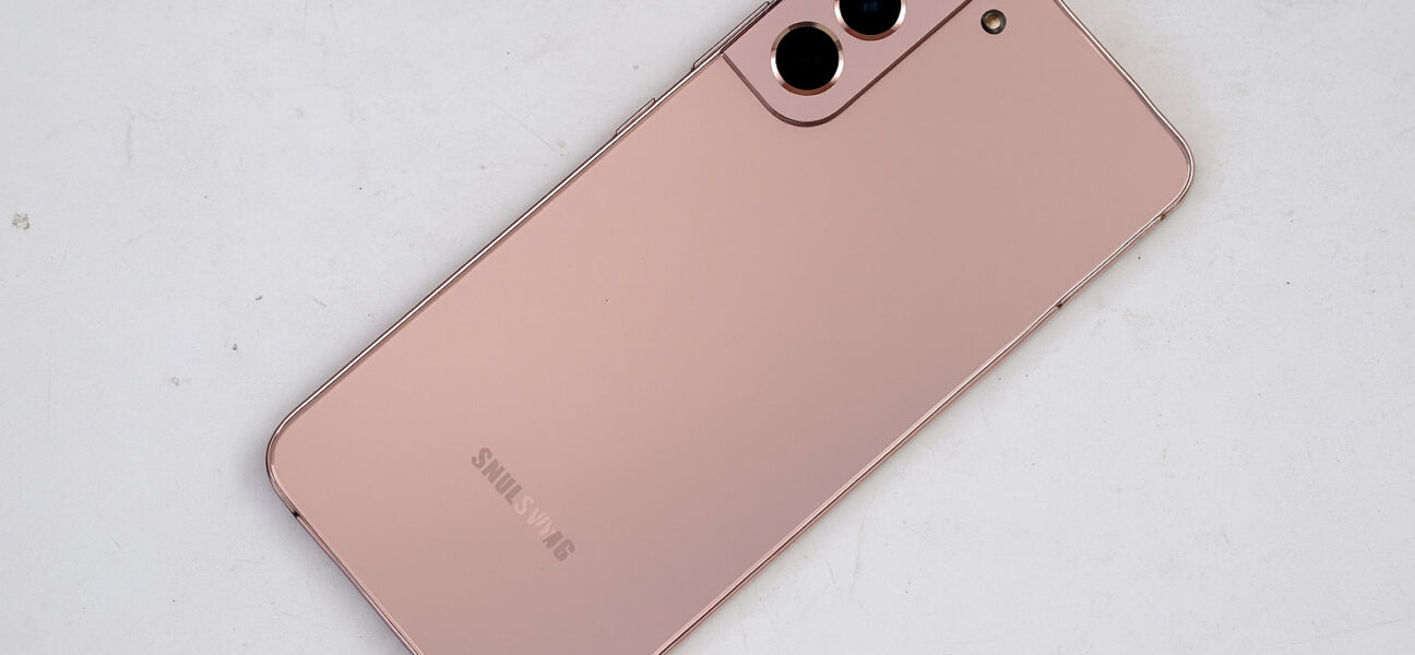 Samsung Galaxy pametni telefon v rožnato-zlati barvi, položen diagonalno na belo, rahlo umazano mizo, s tremi zadnjimi kamerami in mat zaključkom ohišja.