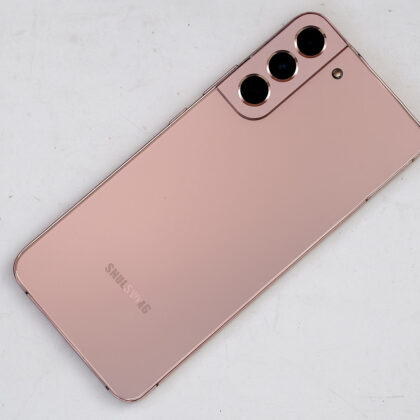 Samsung Galaxy pametni telefon v rožnato-zlati barvi, položen diagonalno na belo, rahlo umazano mizo, s tremi zadnjimi kamerami in mat zaključkom ohišja.