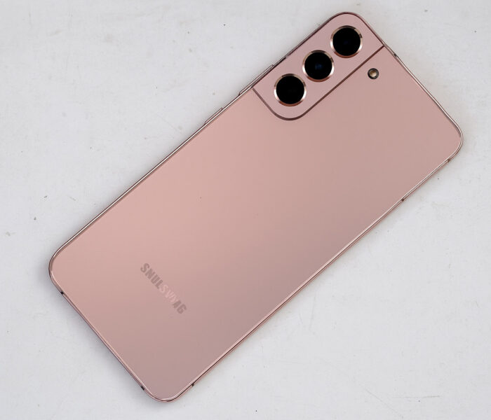 Samsung Galaxy pametni telefon v rožnato-zlati barvi, položen diagonalno na belo, rahlo umazano mizo, s tremi zadnjimi kamerami in mat zaključkom ohišja.