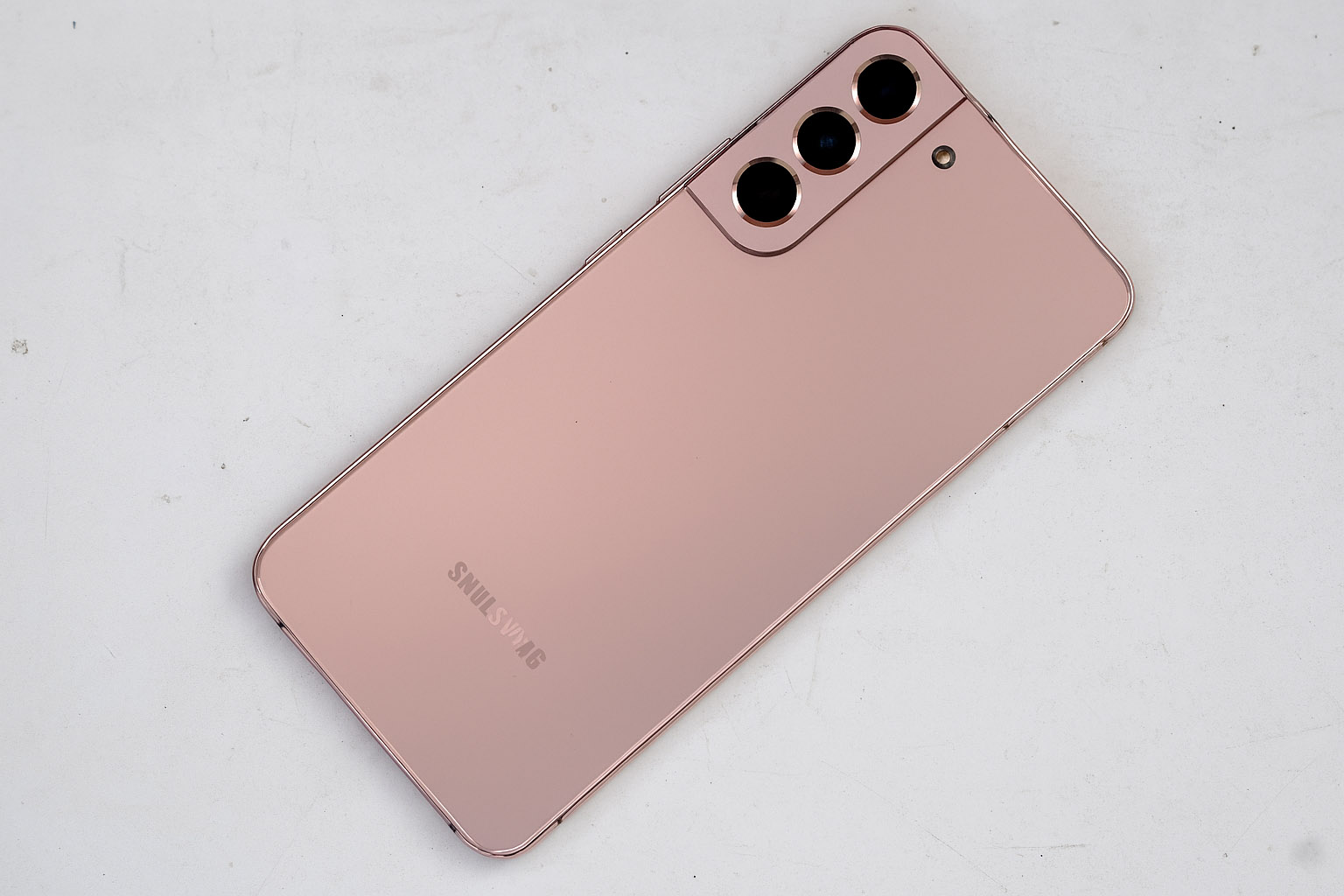 Samsung Galaxy pametni telefon v rožnato-zlati barvi, položen diagonalno na belo, rahlo umazano mizo, s tremi zadnjimi kamerami in mat zaključkom ohišja.